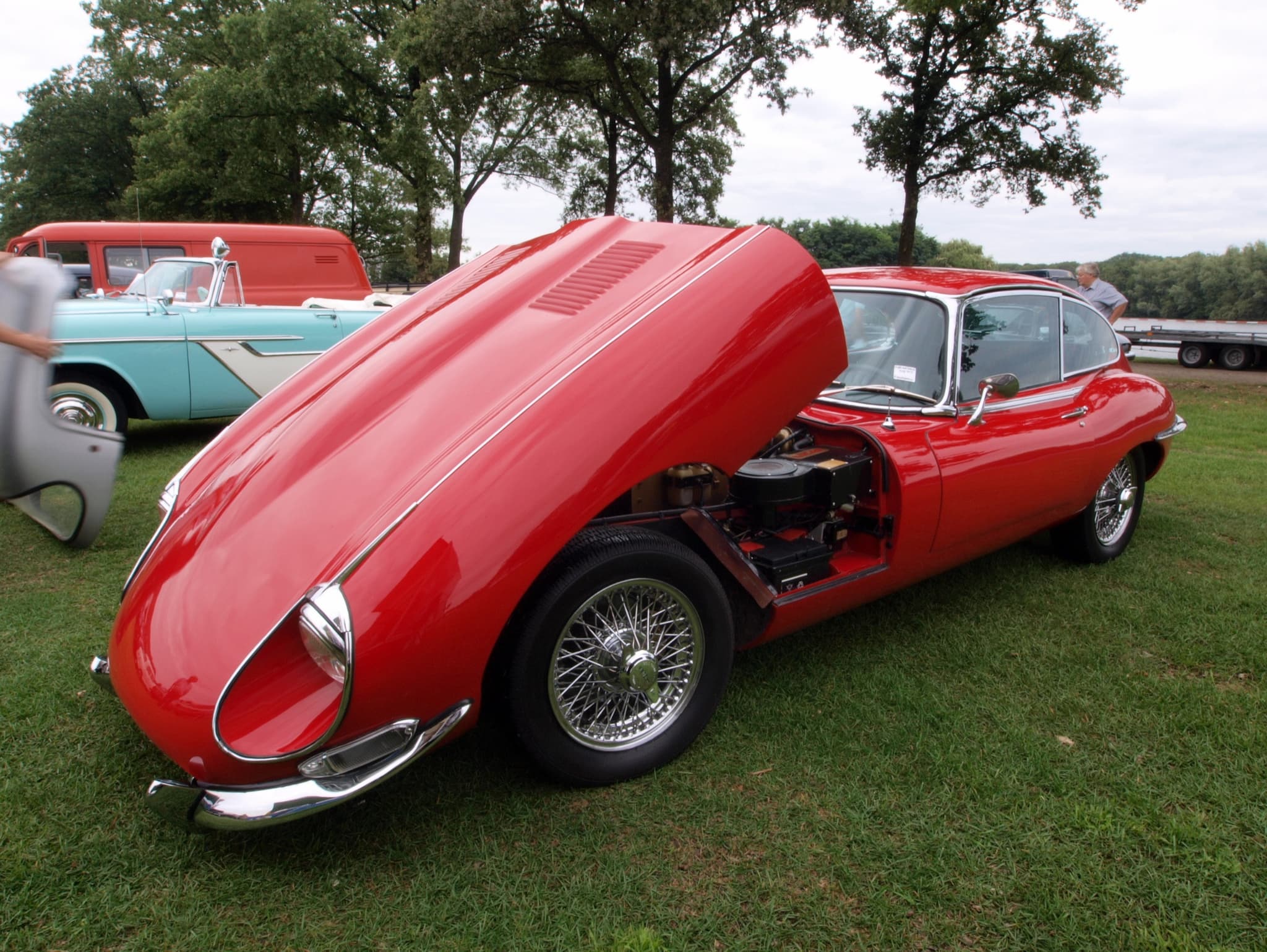 A red E-Type 2+2 with its whole bonnet tipped forward to show the engine, on a grass show field. Opens a larger view