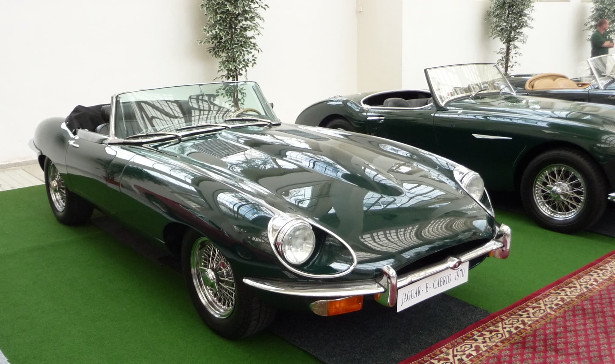 A dark green E-Type roadster with its hood folded, on green display carpet in a white hall.
