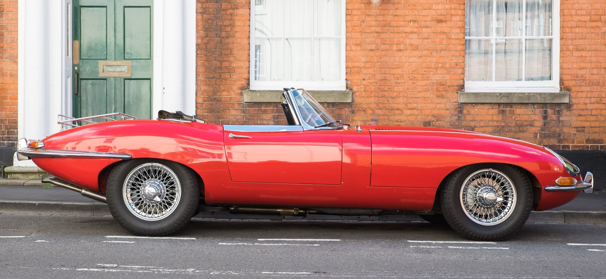 A red E-Type roadster with a chrome boot rack, seen in profile at the kerb in front of a brick house with a green door. Opens a larger view