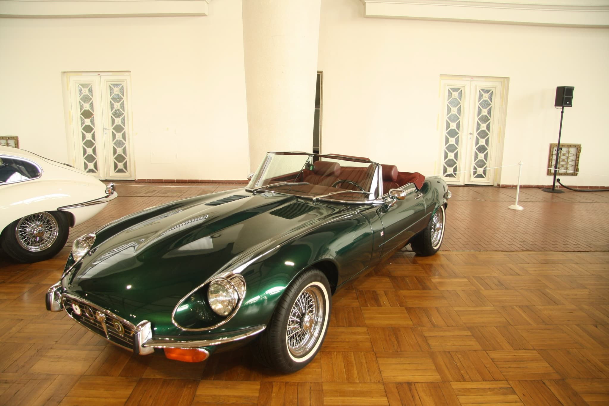 A dark green E-Type roadster with red seats and whitewall tyres on a parquet floor in a white hall.
