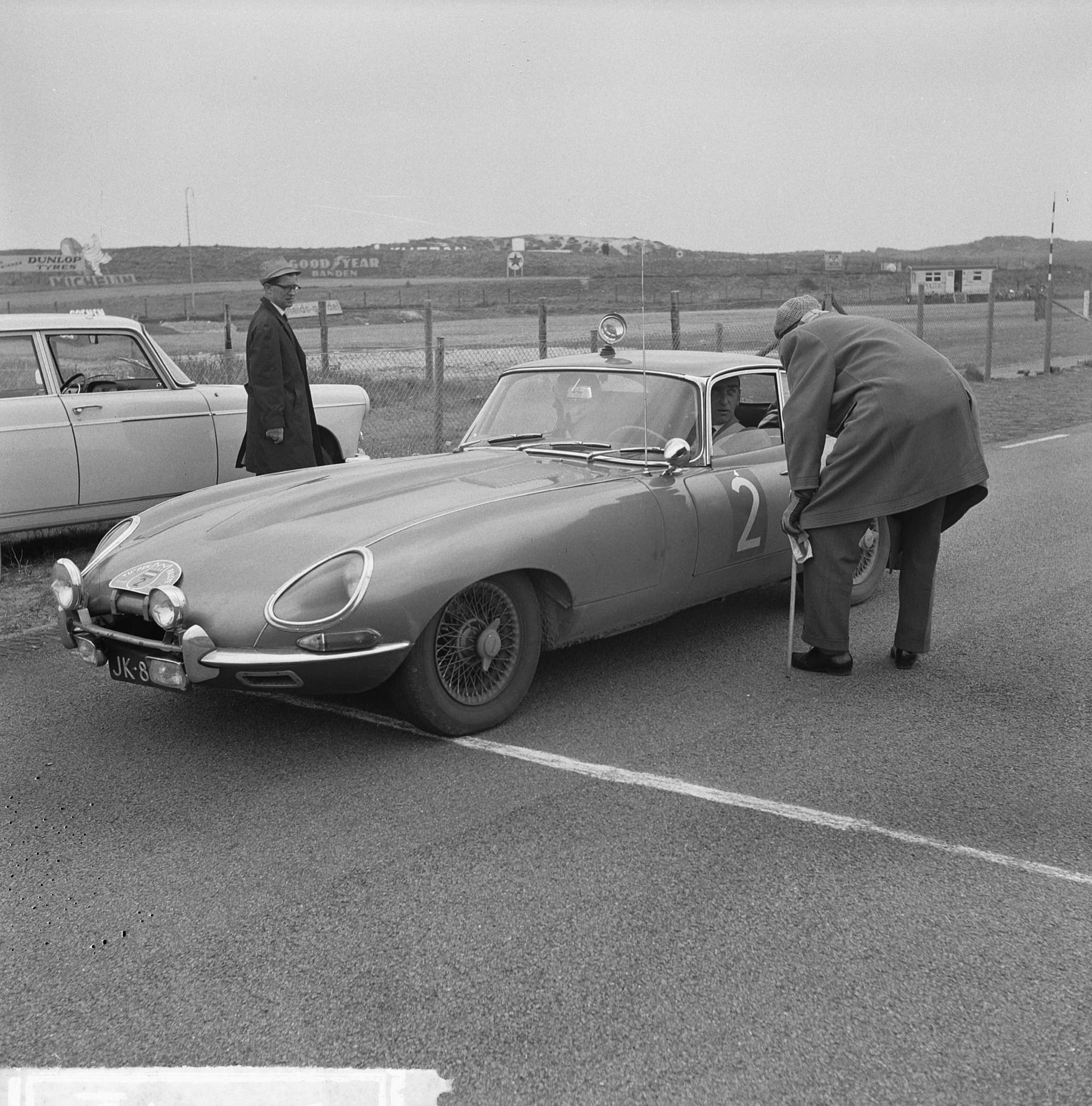 A black-and-white photograph of an E-Type coupé with rally number 2 and a roof lamp, two men in coats beside it on a circuit apron. Opens a larger view