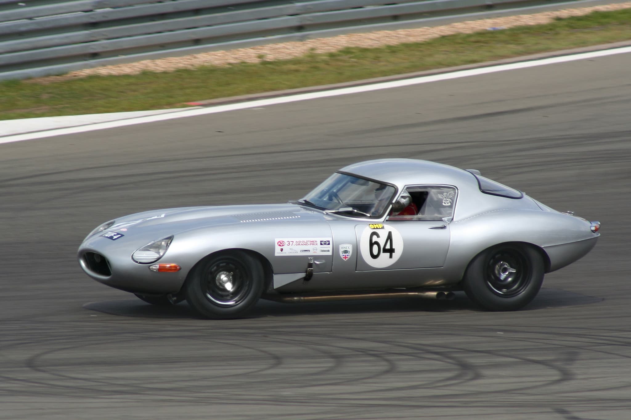 A silver E-Type coupé numbered 64 racing on a circuit.