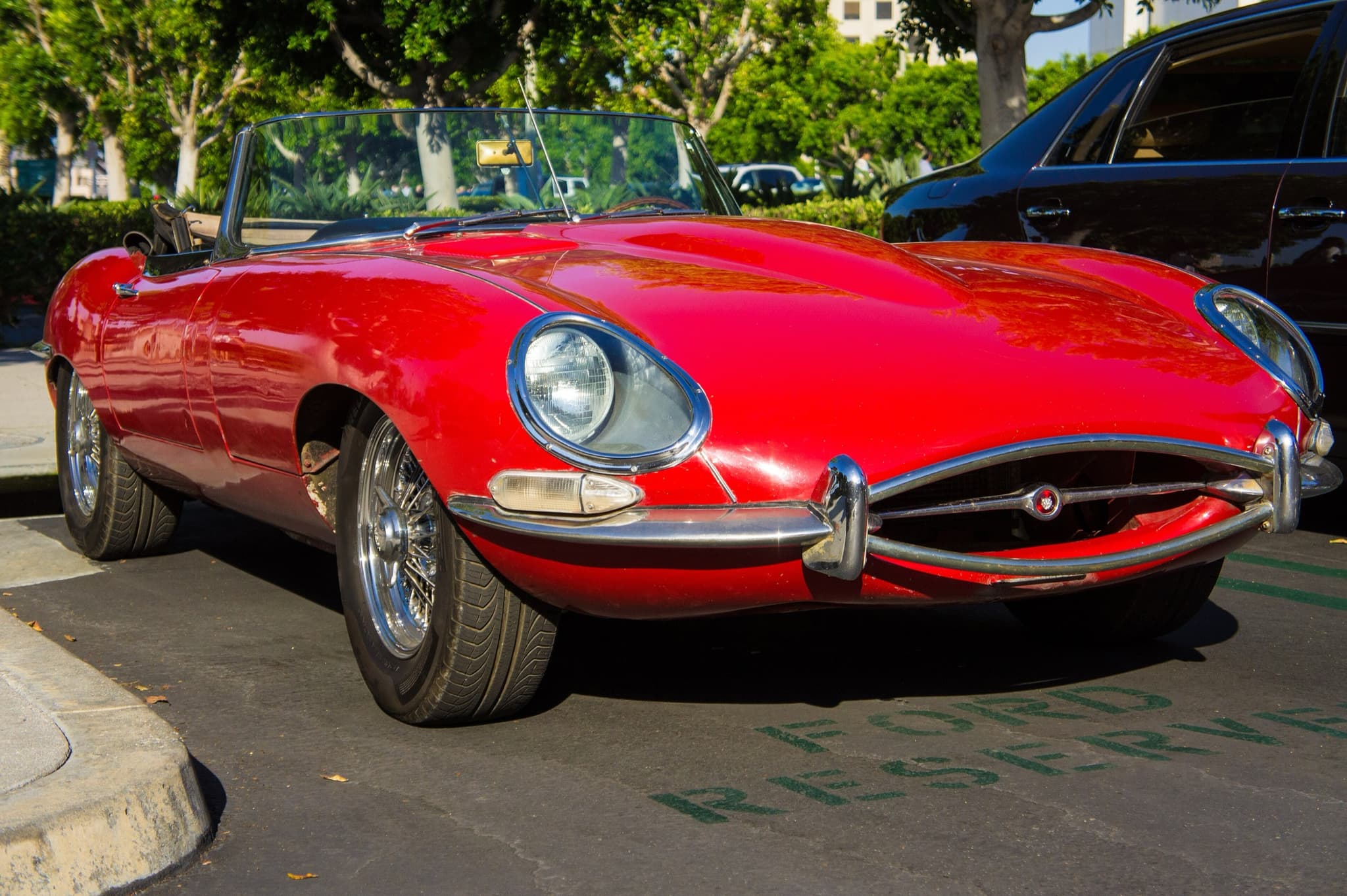 A red E-Type roadster with bubbled paint on its wing and sill, parked in a sunny parking bay.