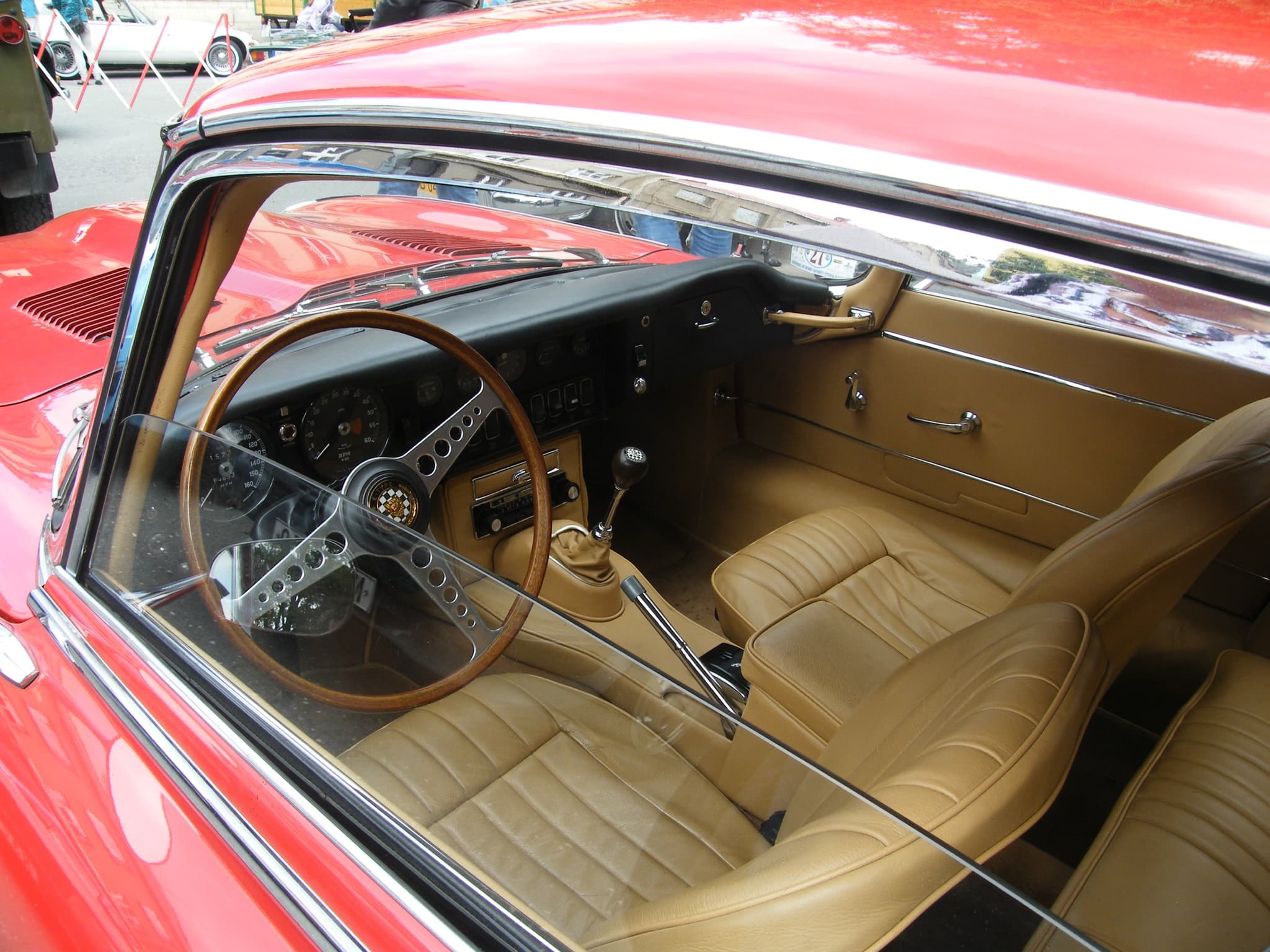 The interior of a red E-Type 2+2 through its window: tan seats, a wood-rim wheel, the dash and the small rear seat.
