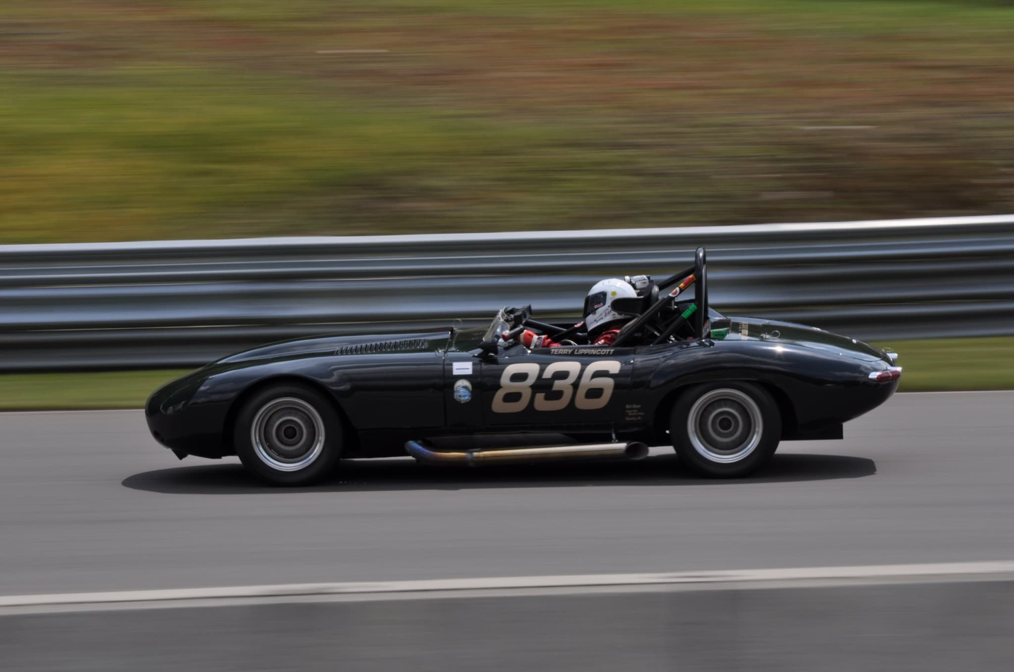 A dark E-Type roadster with a roll hoop and the number 836 racing past a barrier.