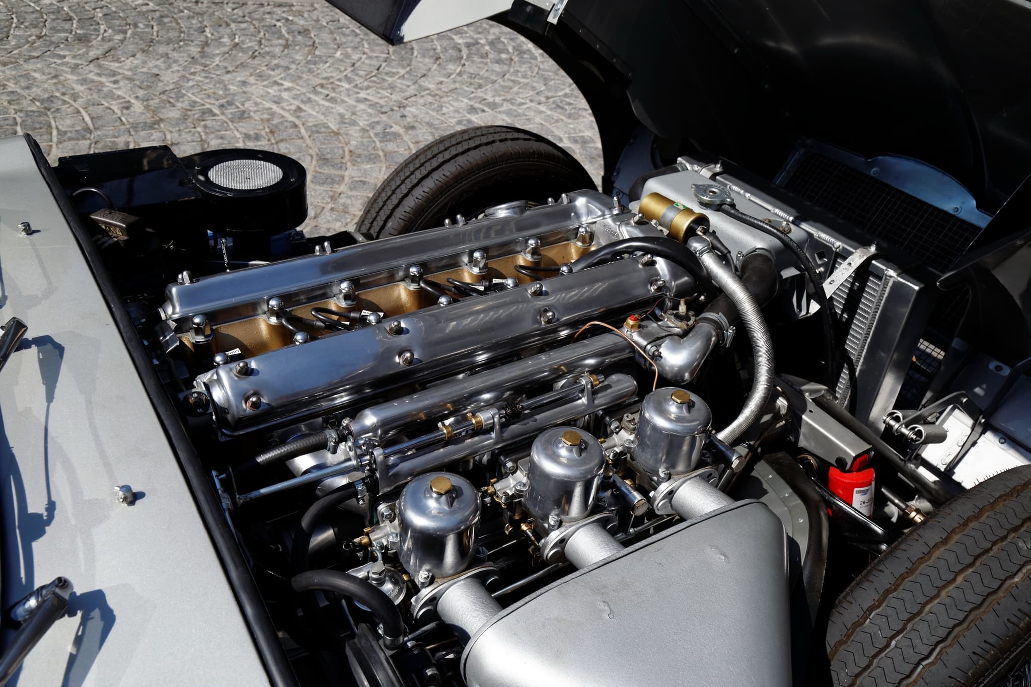 An E-Type's six-cylinder engine with polished cam covers and three carburettors under an aluminium air box.