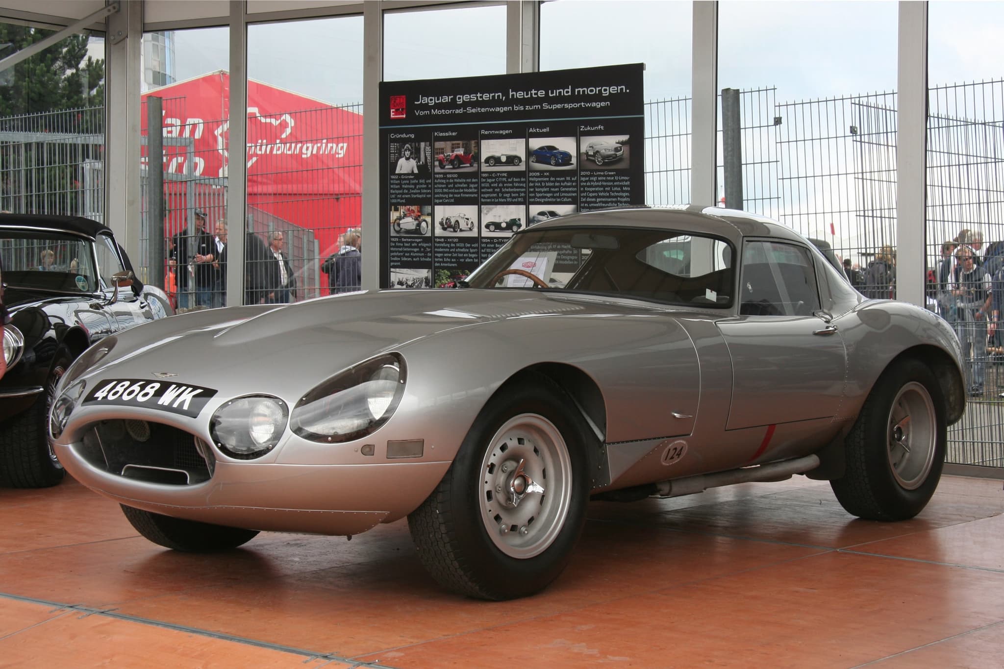 A silver low-drag E-Type with the registration 4868 WK on an orange display floor in a glass pavilion.