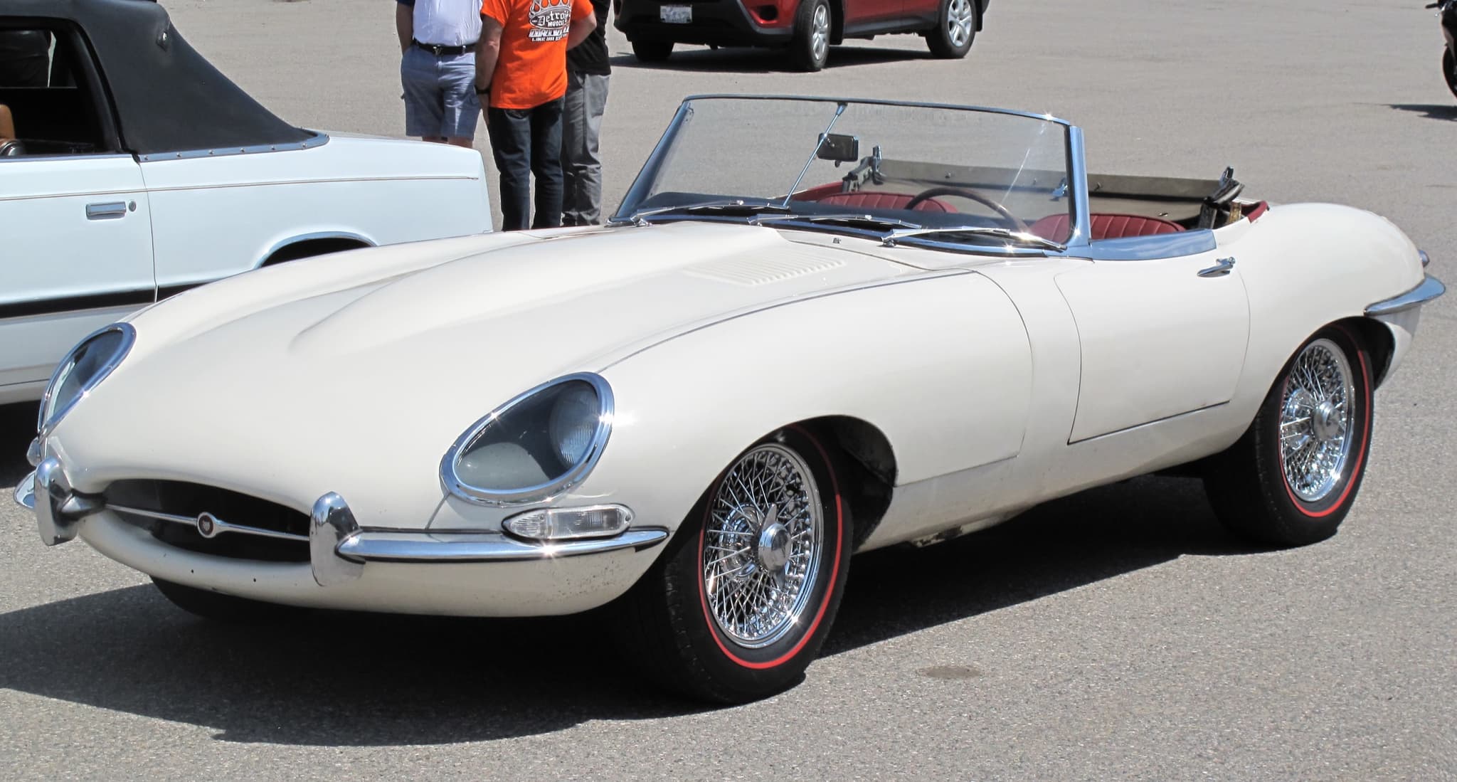 A white E-Type roadster with red seats and red-line tyres on a car park, seen from the front three-quarter.