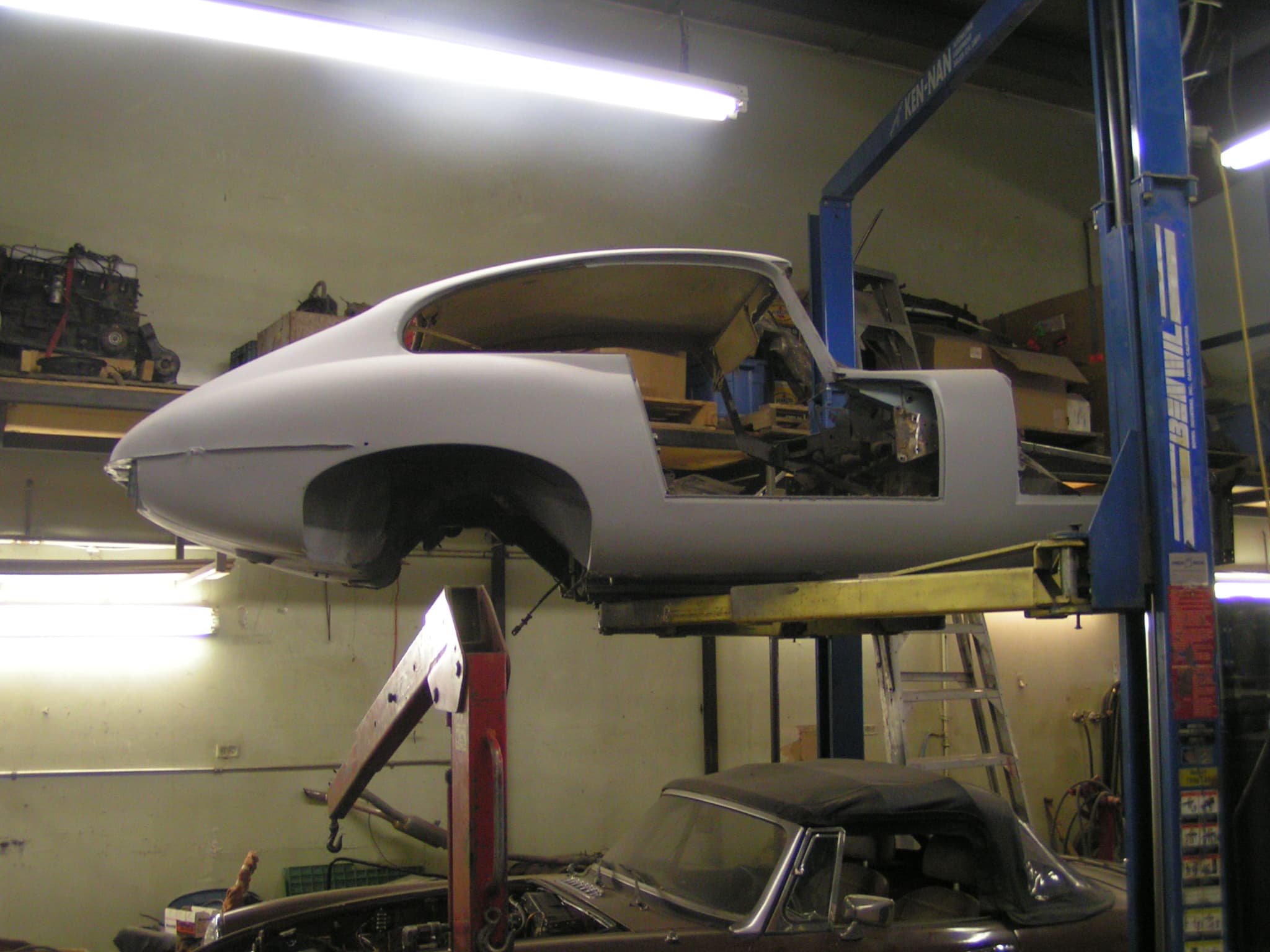 A primed E-Type coupé body shell raised on a two-post lift in a workshop.