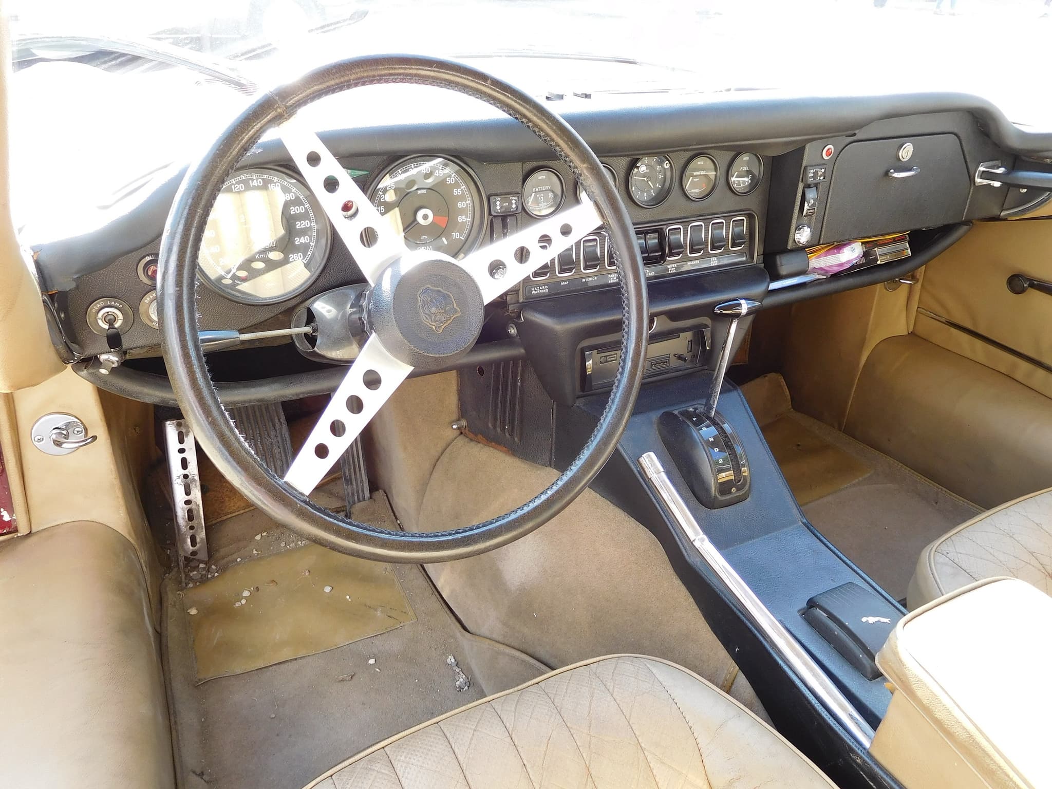 The driving seat of a maroon E-Type 2+2 through its open door: a leather-rim wheel, dials, rocker switches and an automatic gear selector.