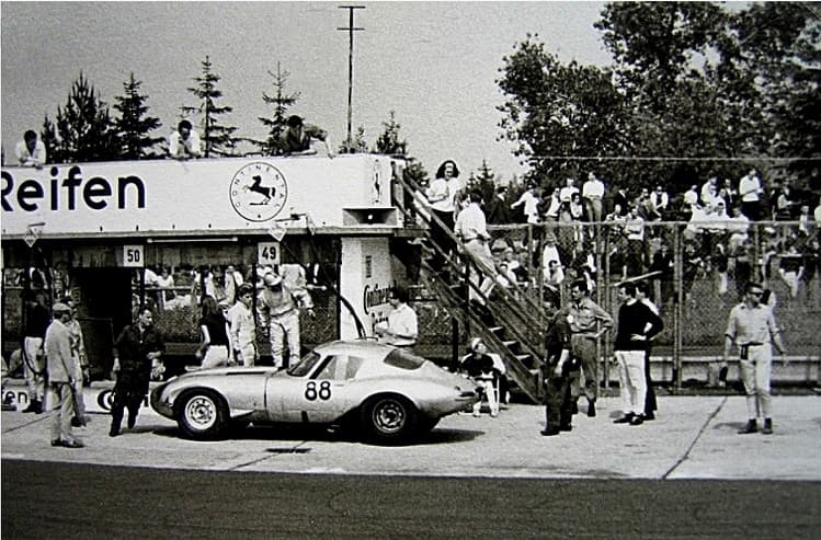 A black-and-white photograph of a silver low-drag E-Type coupé numbered 88 at a pit stop, surrounded by mechanics, with a crowded grandstand behind.
