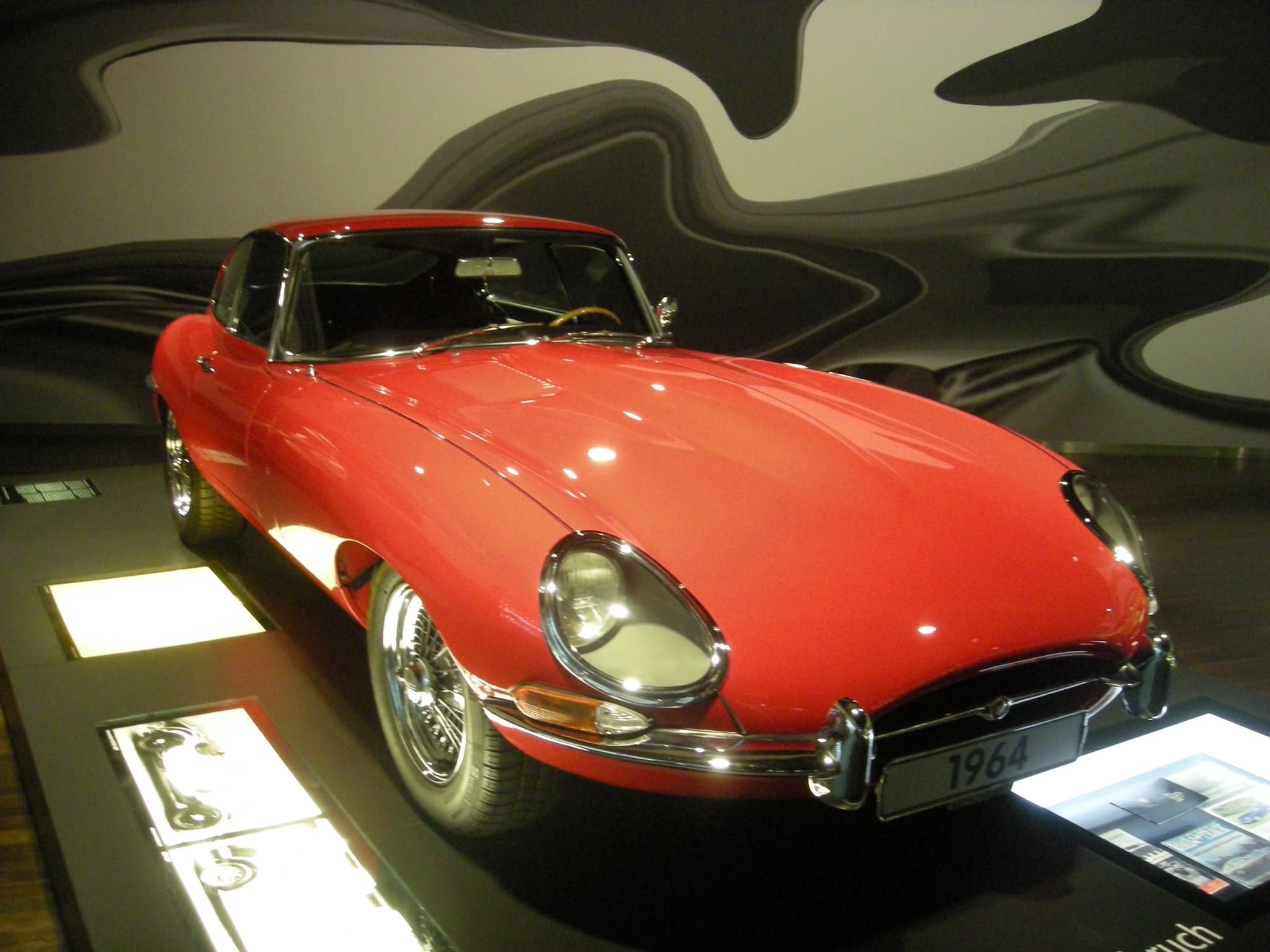 A red E-Type coupé on a dark plinth in a dimly lit exhibition hall.
