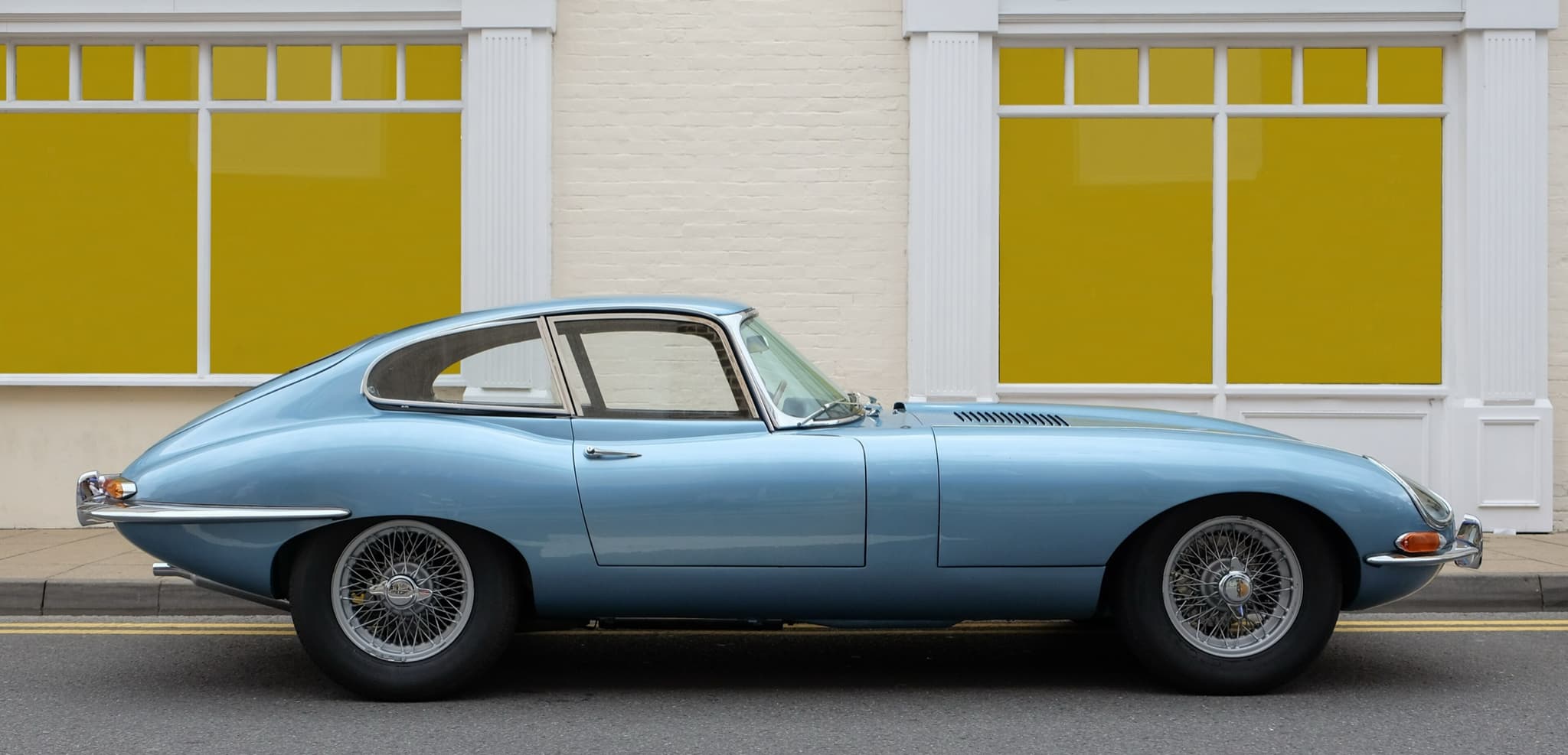A pale blue E-Type coupé in profile at the kerb, in front of a brick shopfront with yellow windows.