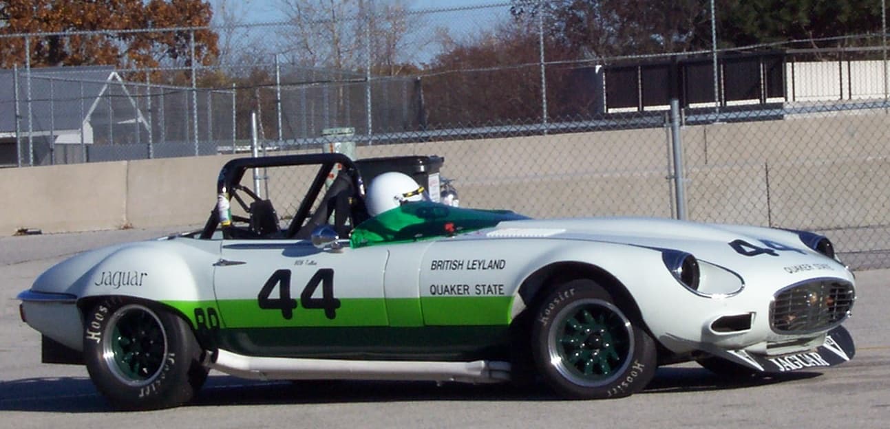 A white and green E-Type V12 race car numbered 44 on a circuit, its helmeted driver in the cockpit.