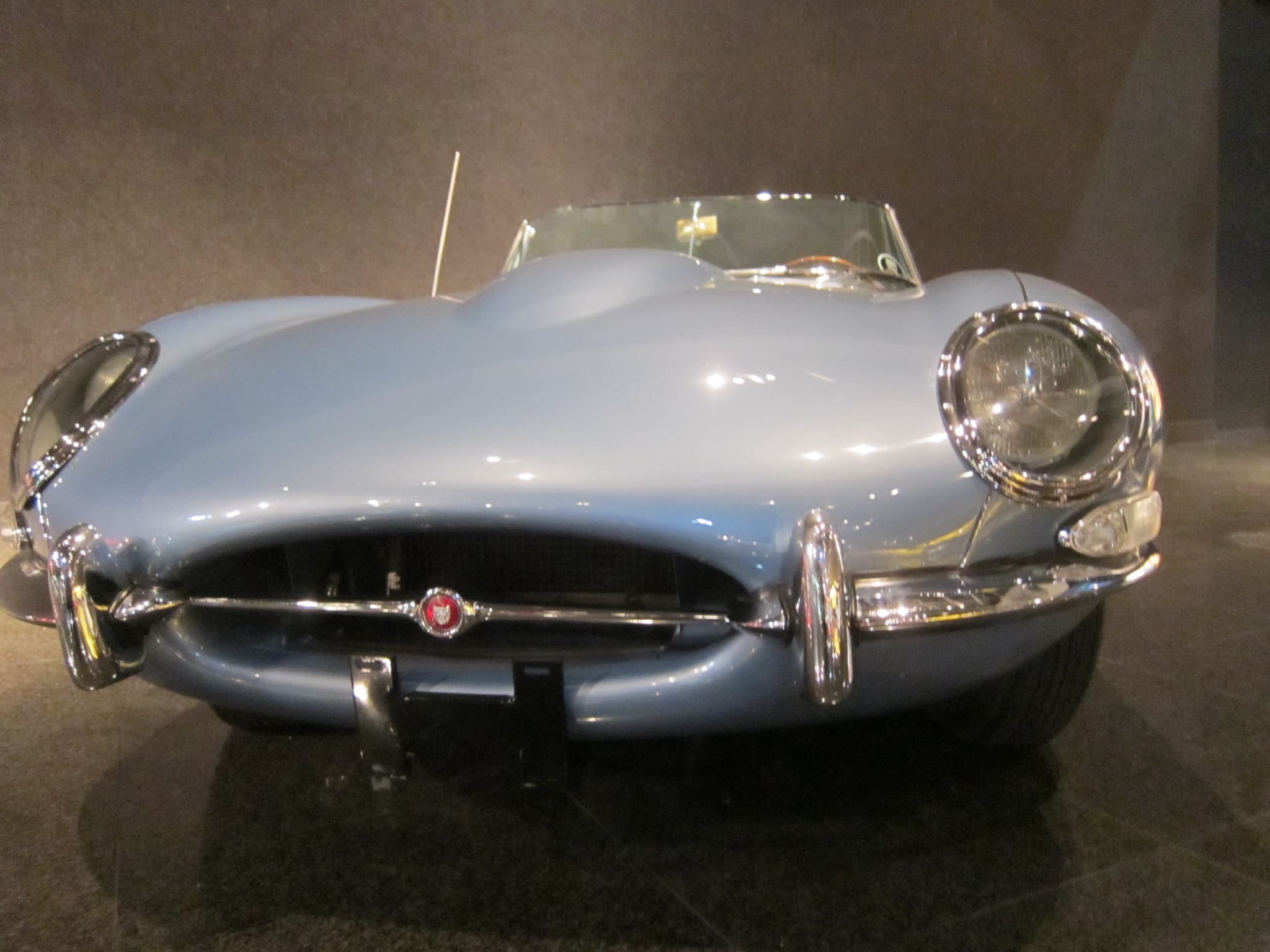 A pale blue E-Type roadster seen head-on in a dark museum hall, showing its glass-covered headlamps and small oval air intake.