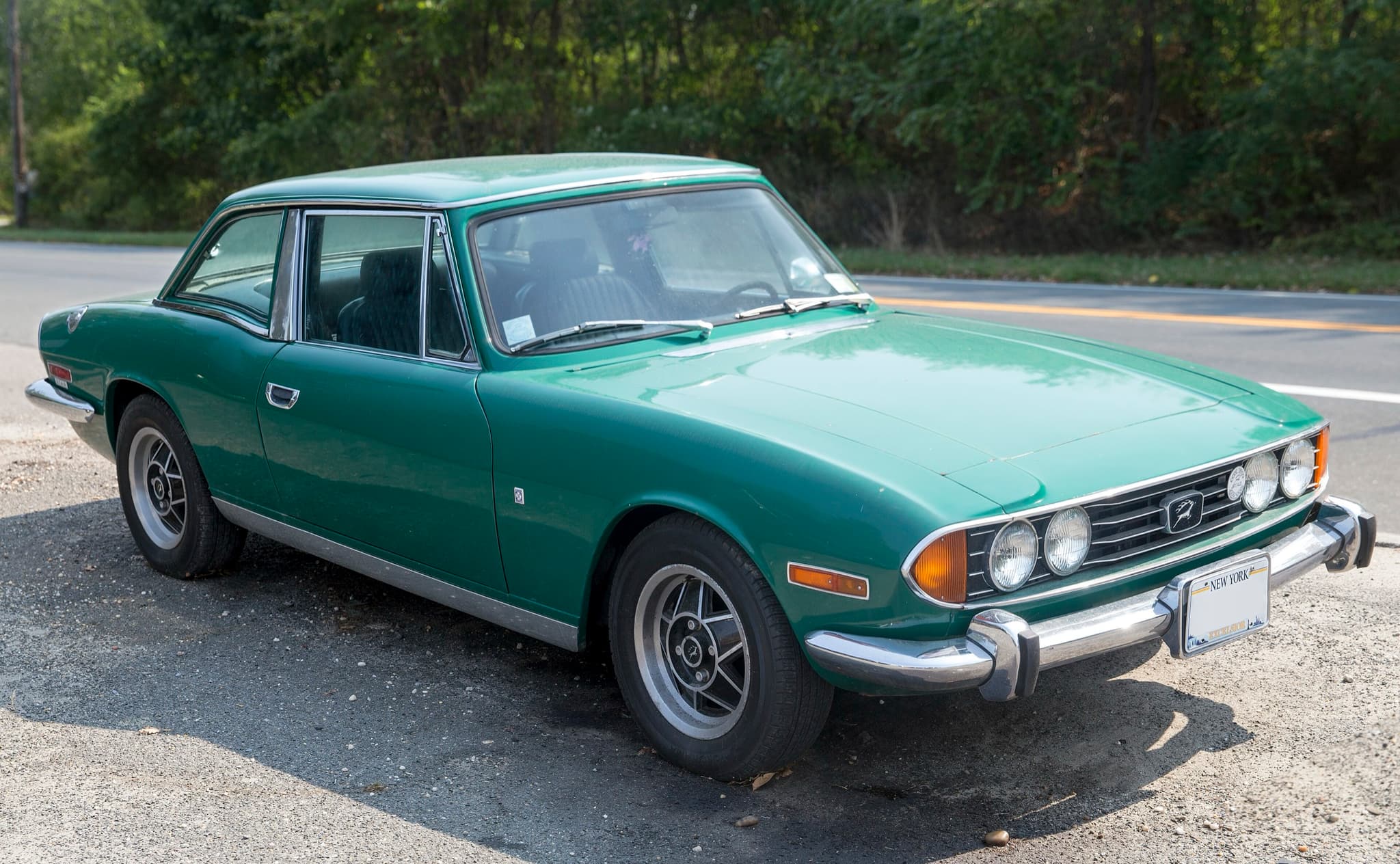 A green Triumph Stag with its hardtop parked on gravel beside a road.