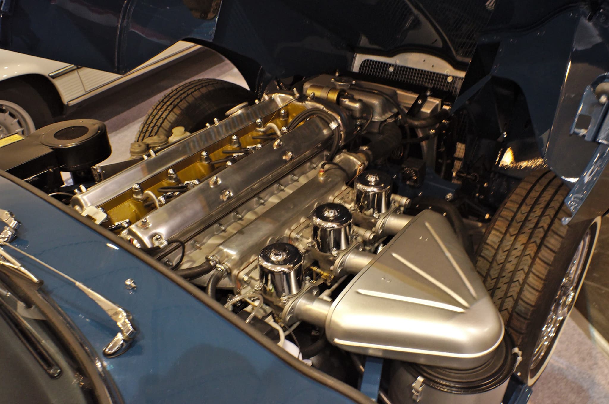 The engine bay of a blue E-Type with its bonnet tipped forward: a six-cylinder engine with three carburettors and an aluminium air box.
