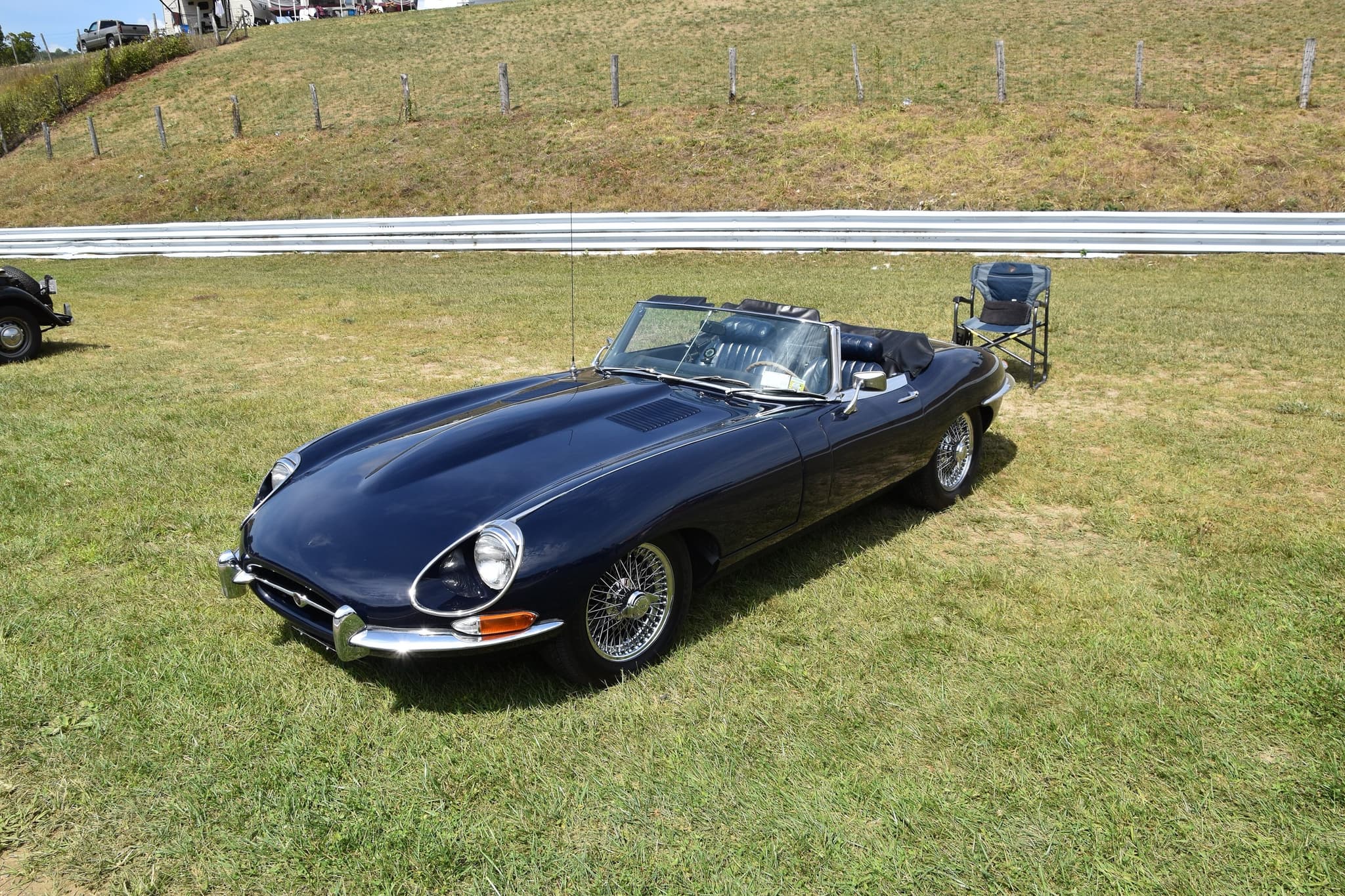 A dark blue E-Type roadster with its hood folded, parked on grass beside a circuit's steel barrier. Opens a larger view