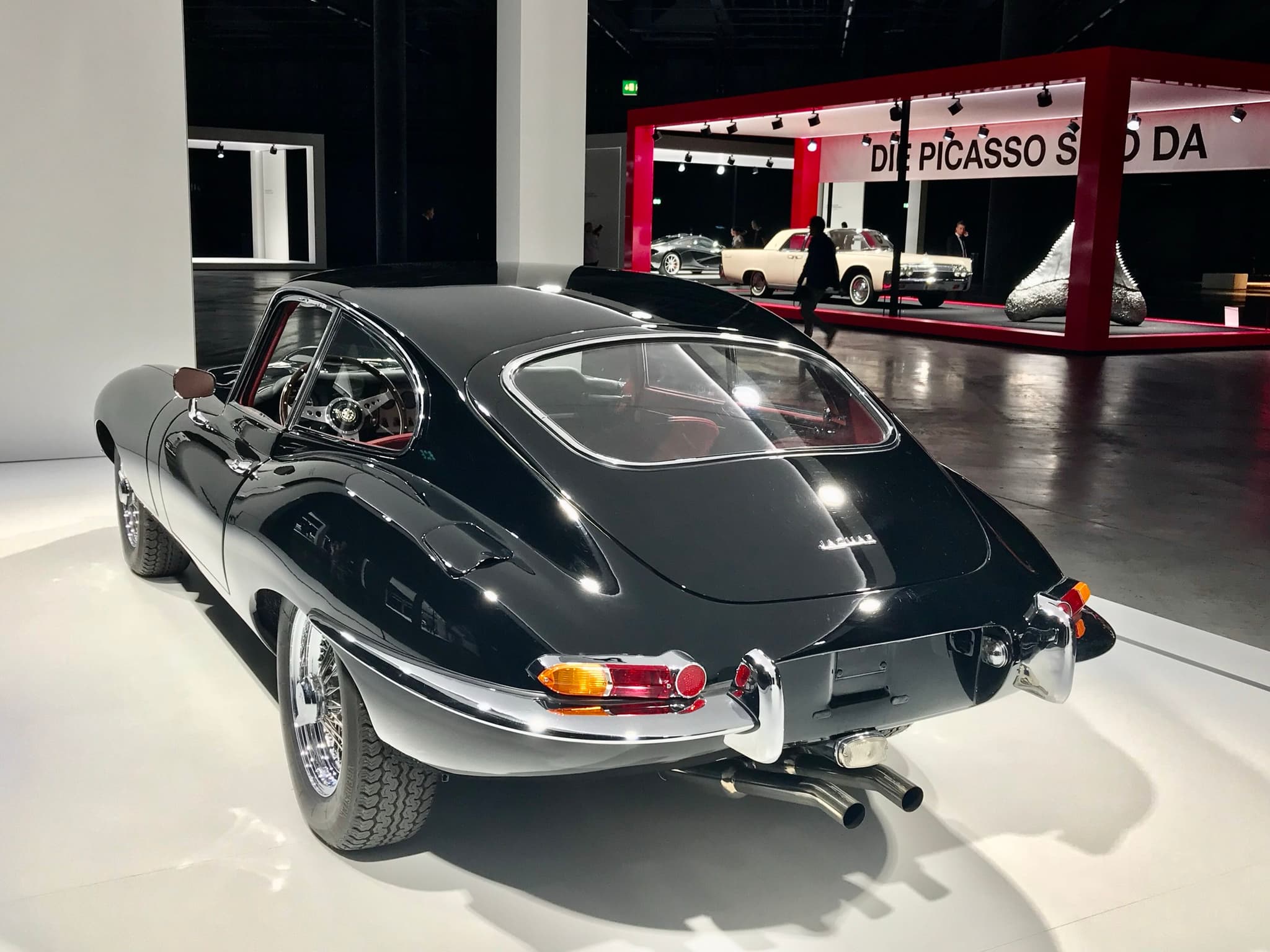 The tail of a black E-Type coupé, showing its side-hinged rear door, small tail lamps and twin exhaust pipes.