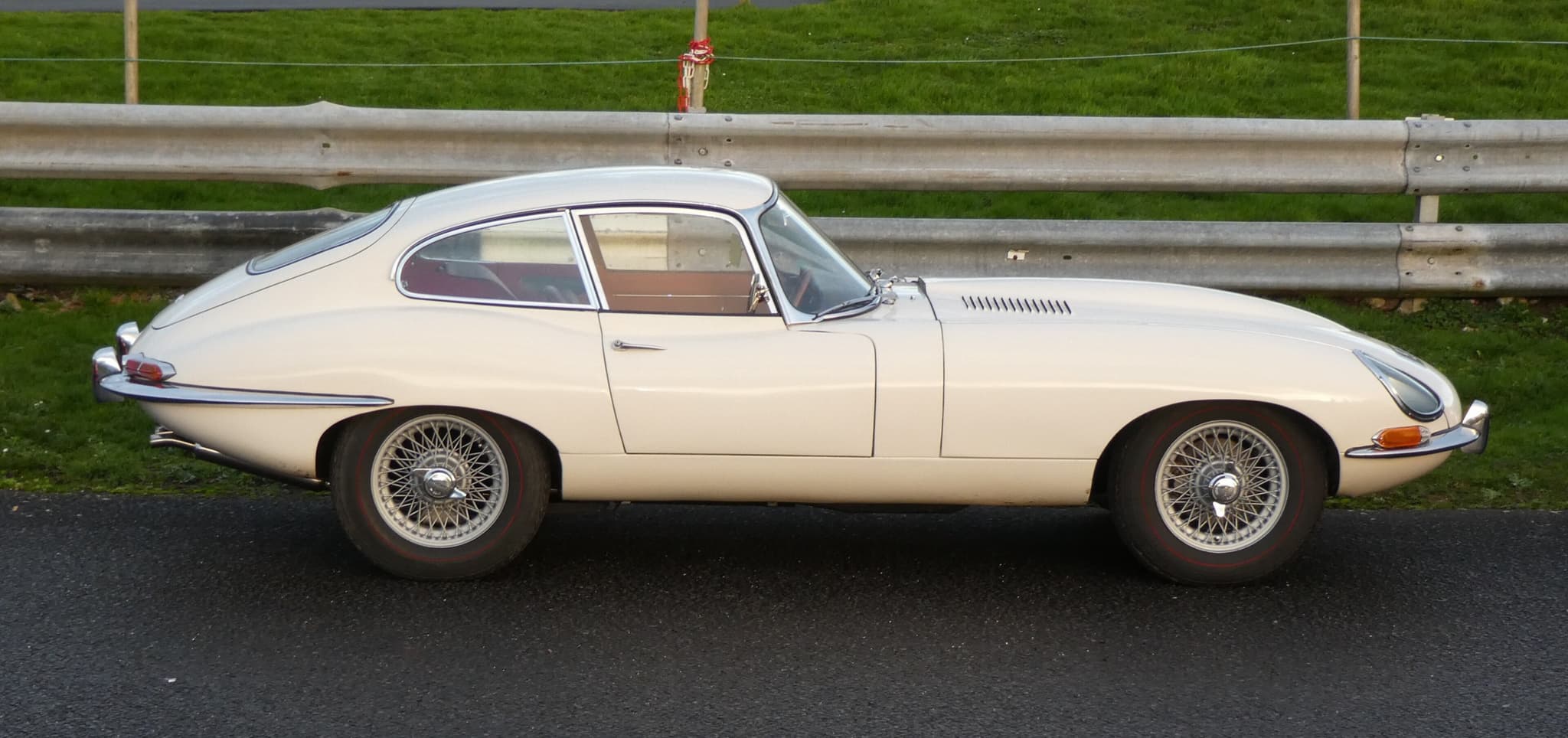 A cream E-Type coupé in profile on a paddock road, in front of a steel barrier and a grass bank. Opens a larger view
