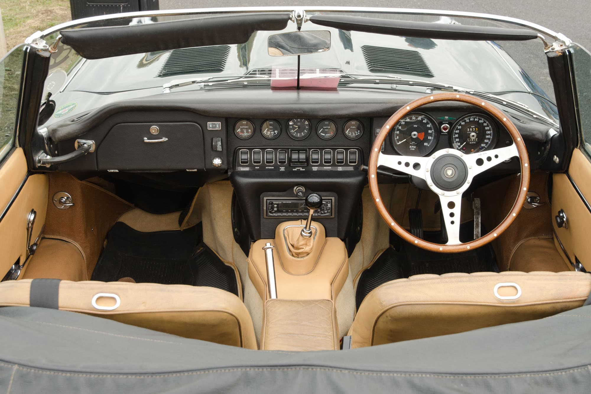 An E-Type roadster's cockpit from above, with tan seats and a row of rocker switches under the centre dials.