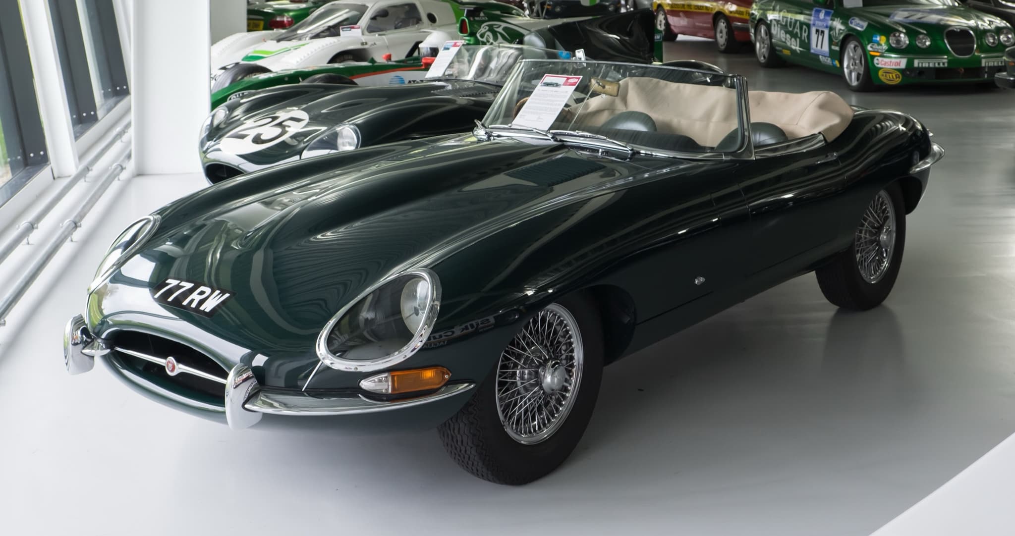 A dark green E-Type roadster with the registration 77 RW and its hood down on a white museum floor.