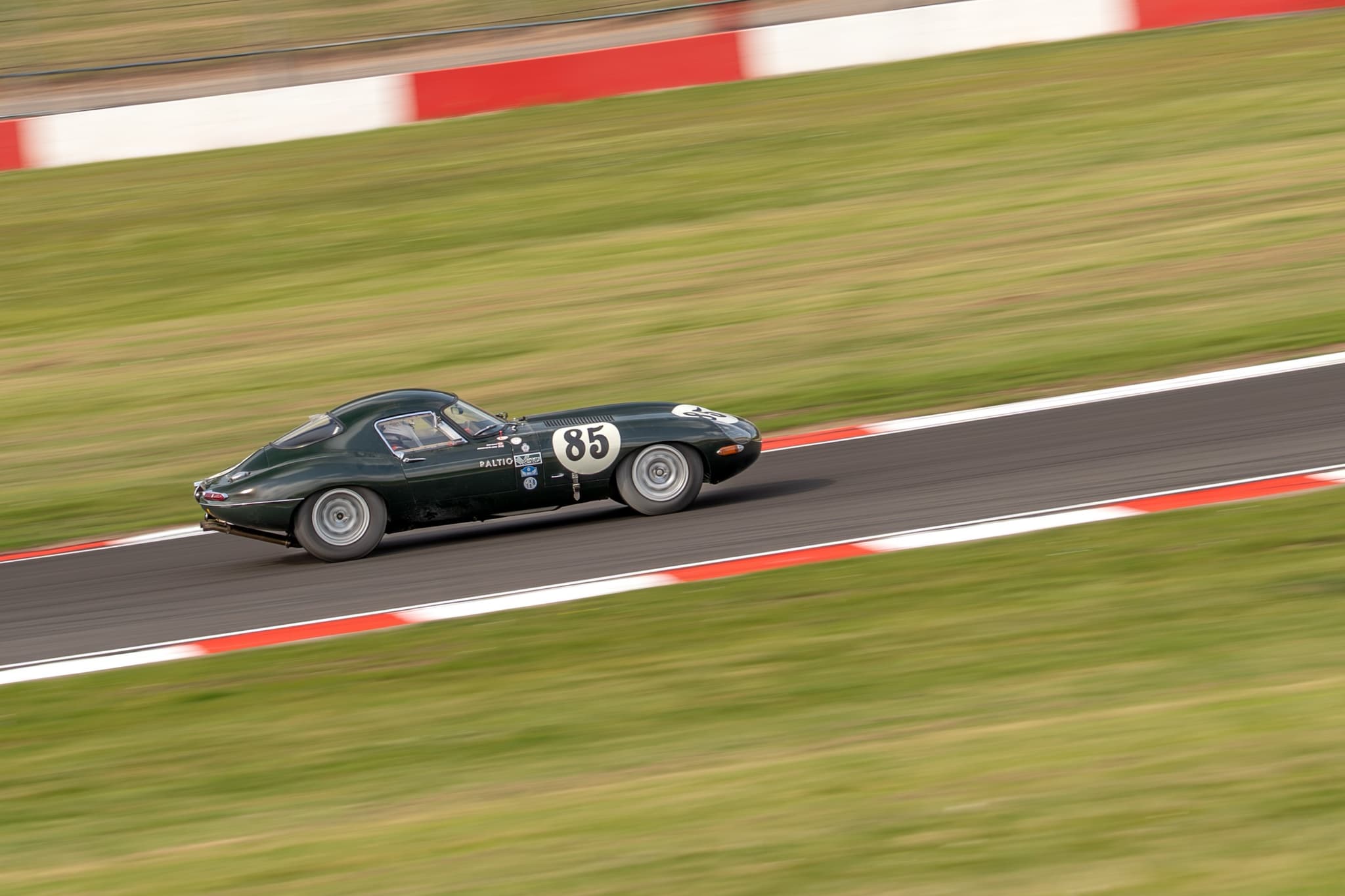 A dark green E-Type coupé numbered 85 racing between red and white kerbs.