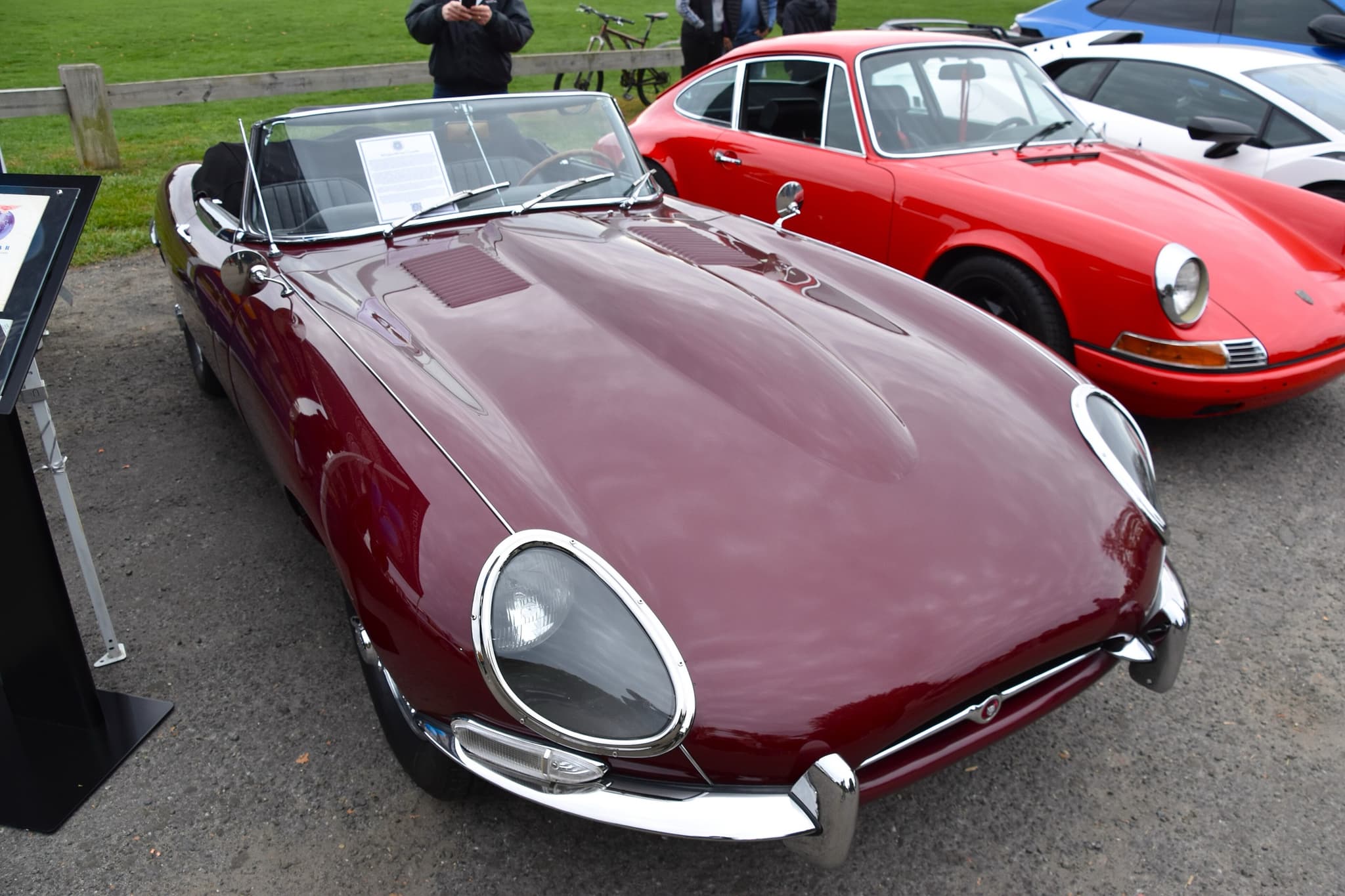 A maroon E-Type roadster seen from above its long bonnet, parked on gravel beside a red Porsche 911.