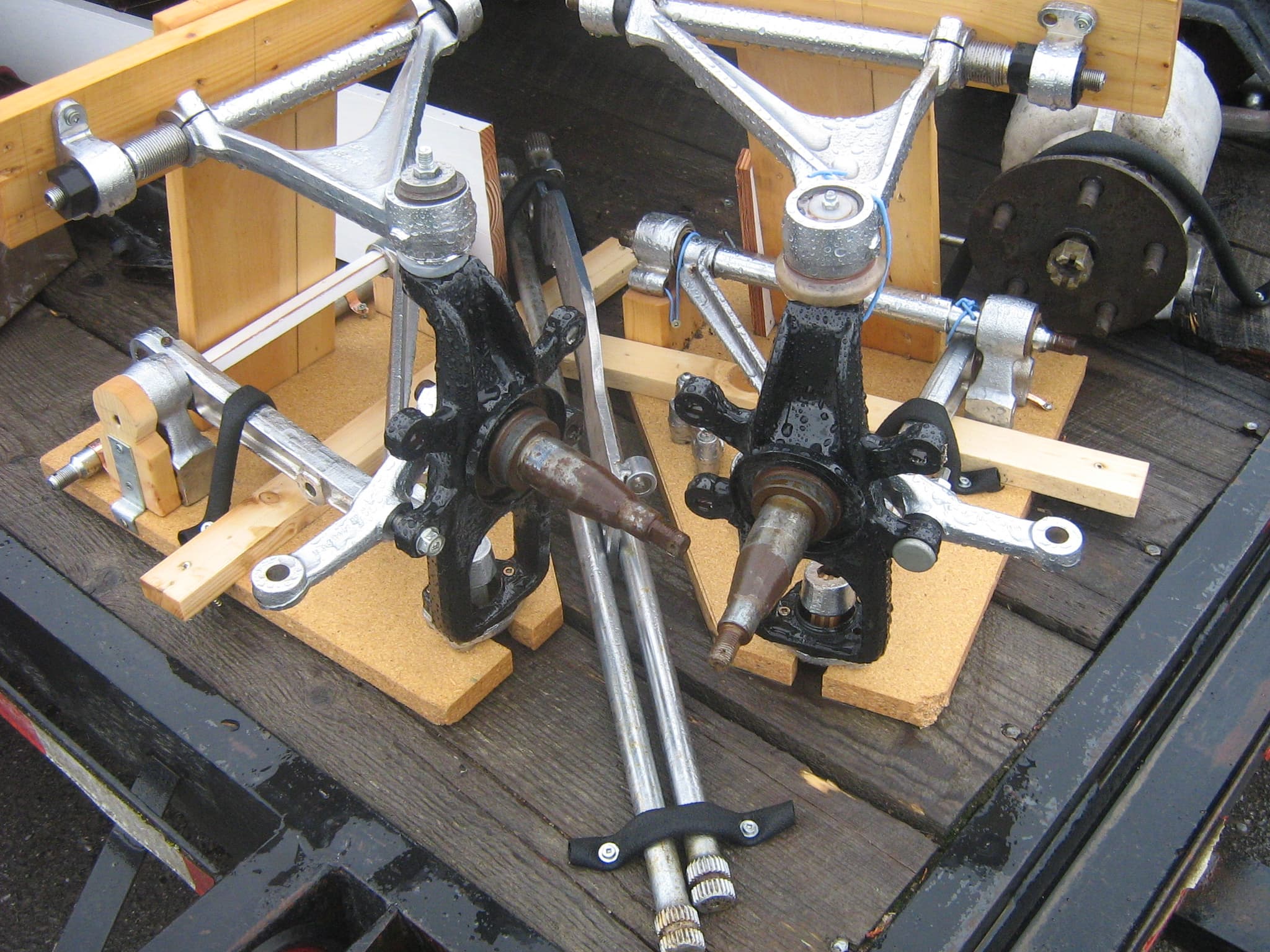 E-Type front suspension parts laid on a trailer: silver wishbones, stub-axle carriers and two long torsion bars.