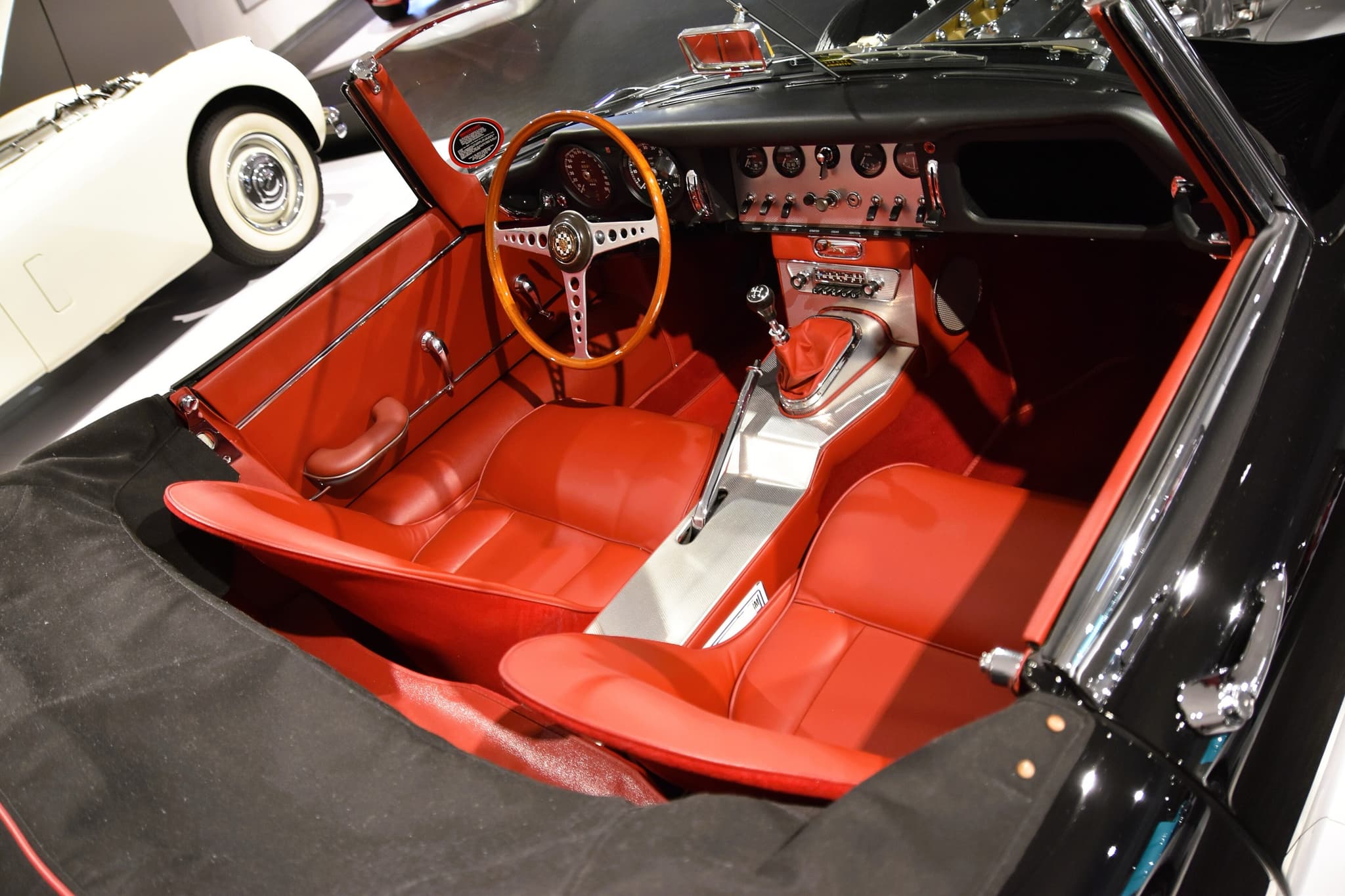 An early E-Type roadster's cockpit from above, with red leather seats, an aluminium centre panel and console, and a wood-rim wheel on the left.