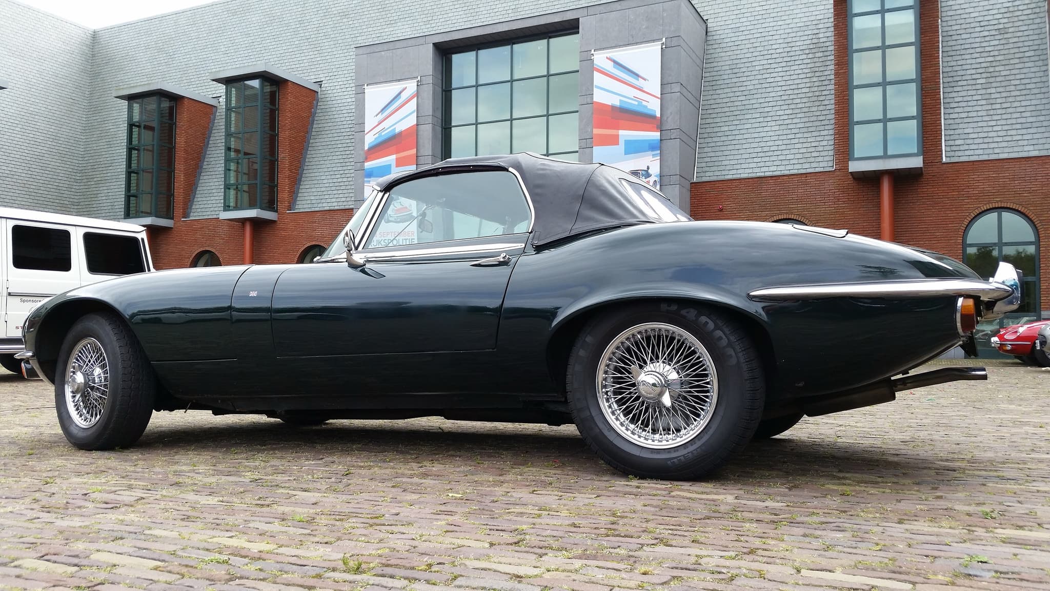 A black E-Type roadster with its hood up and flared wheel arches, in profile on cobbles outside a brick museum.