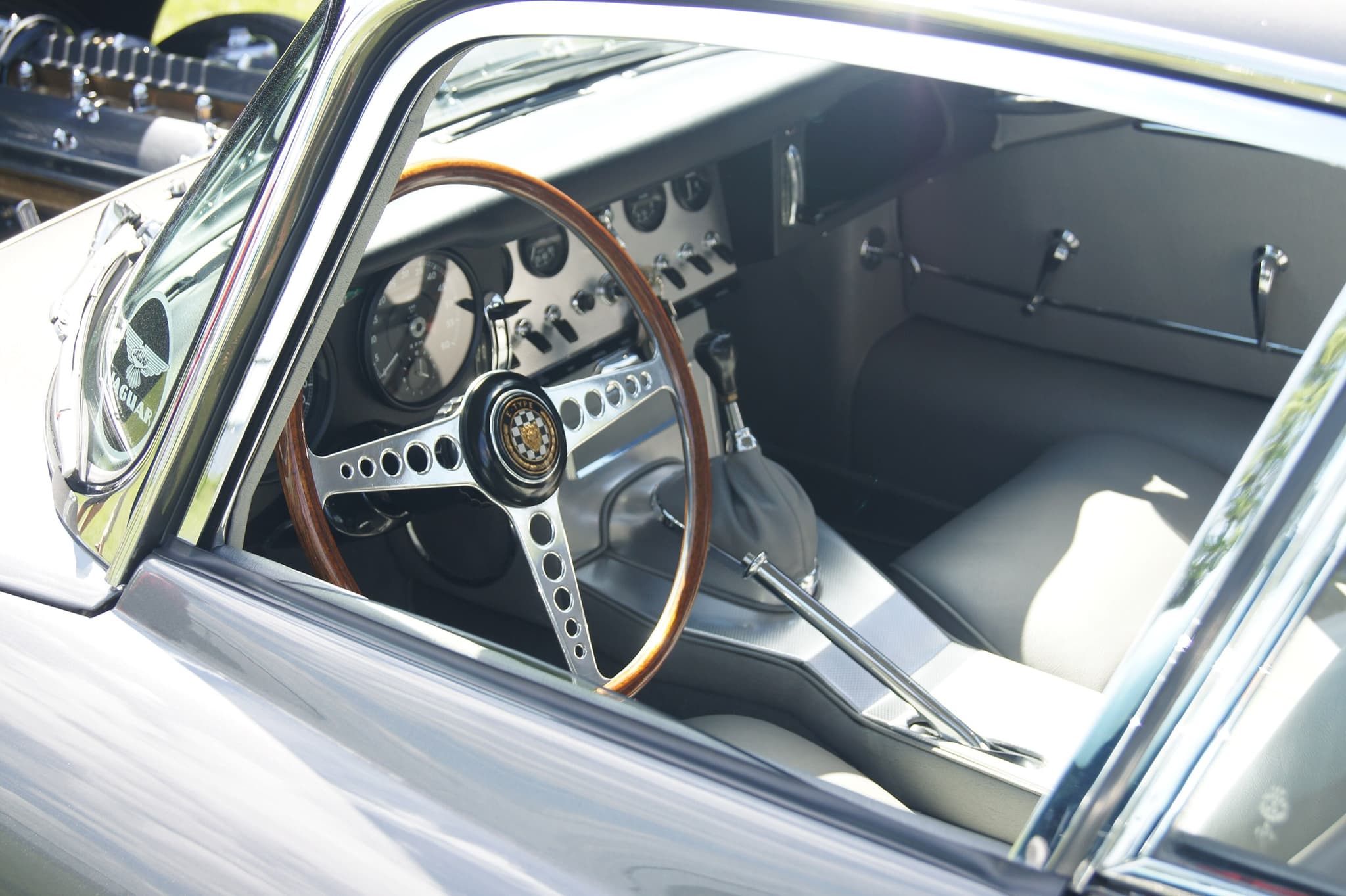 The cockpit of a grey E-Type coupé through its open door: grey leather seats, an aluminium centre panel and a wood-rim wheel on the left.
