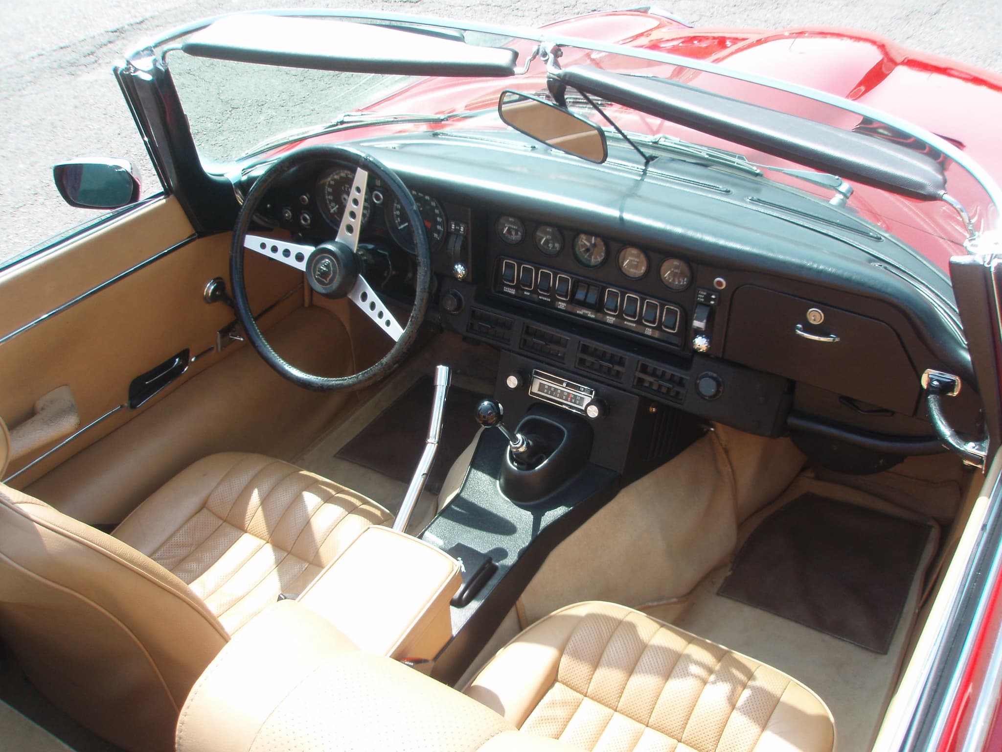 An E-Type roadster's cockpit from above, with tan leather seats, a manual gear lever and a black dash.