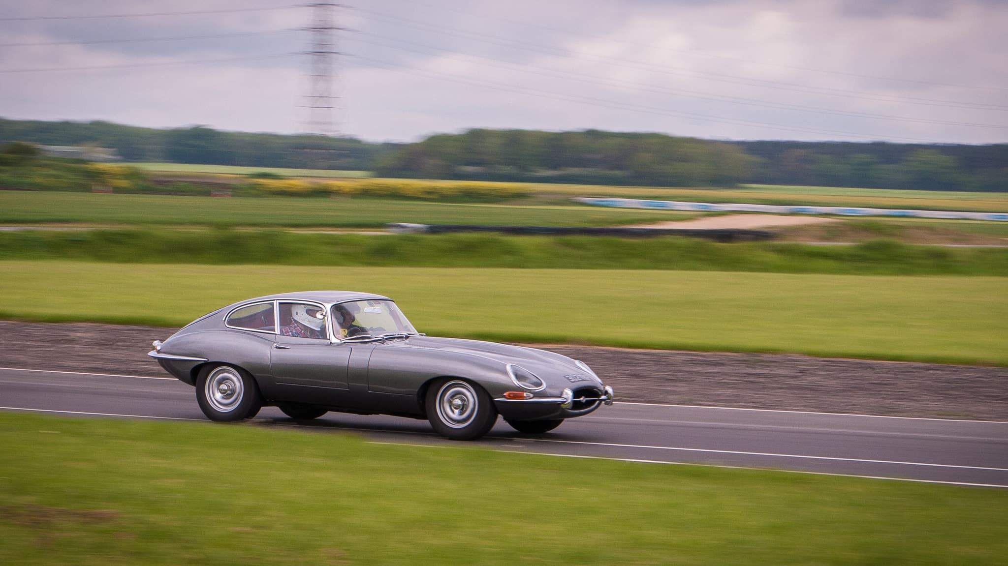 A grey E-Type coupé speeding along a circuit, the grass behind it blurred by the moving camera.