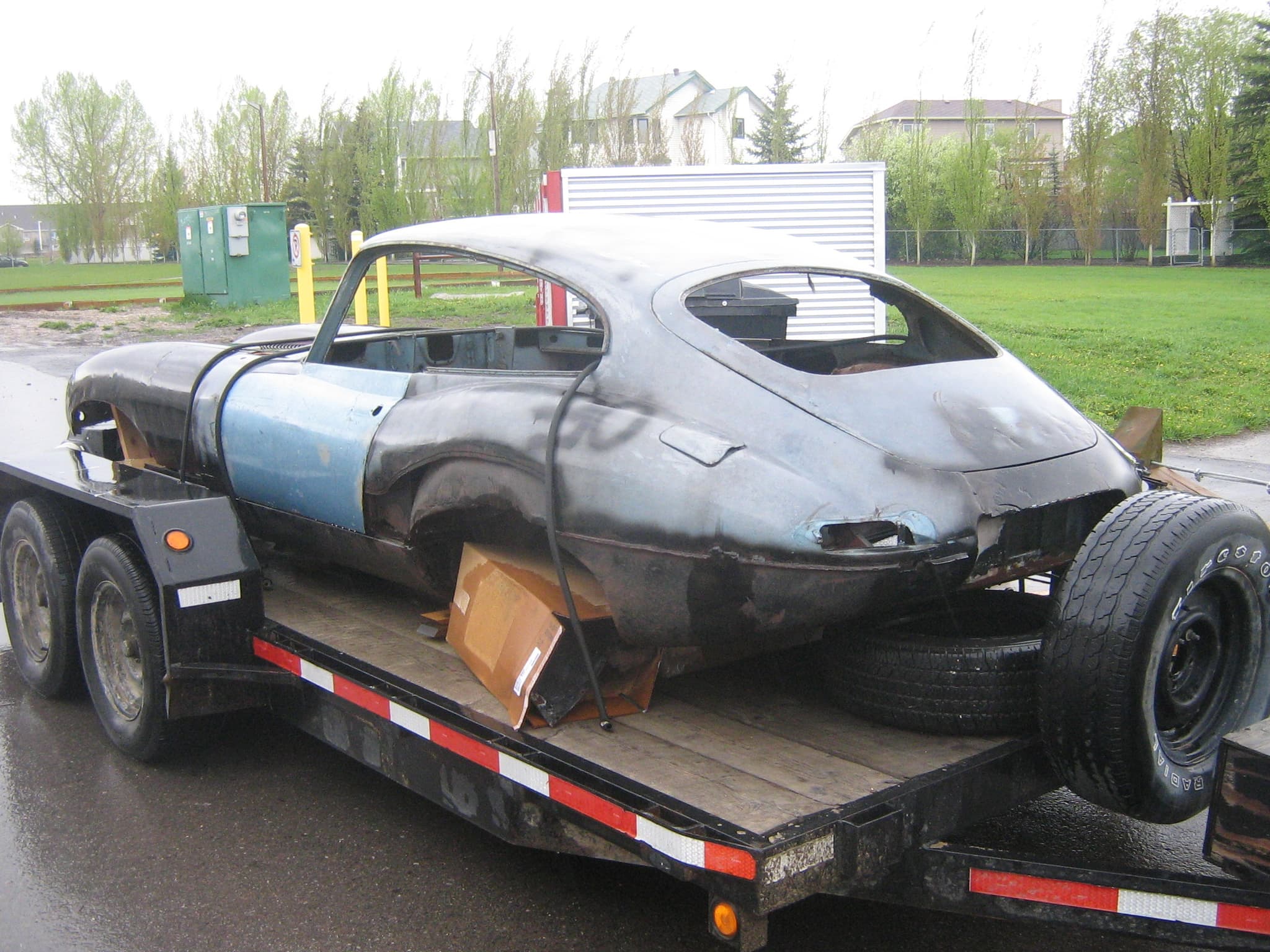 An unrestored E-Type coupé shell with rusted lower panels on a flatbed trailer.