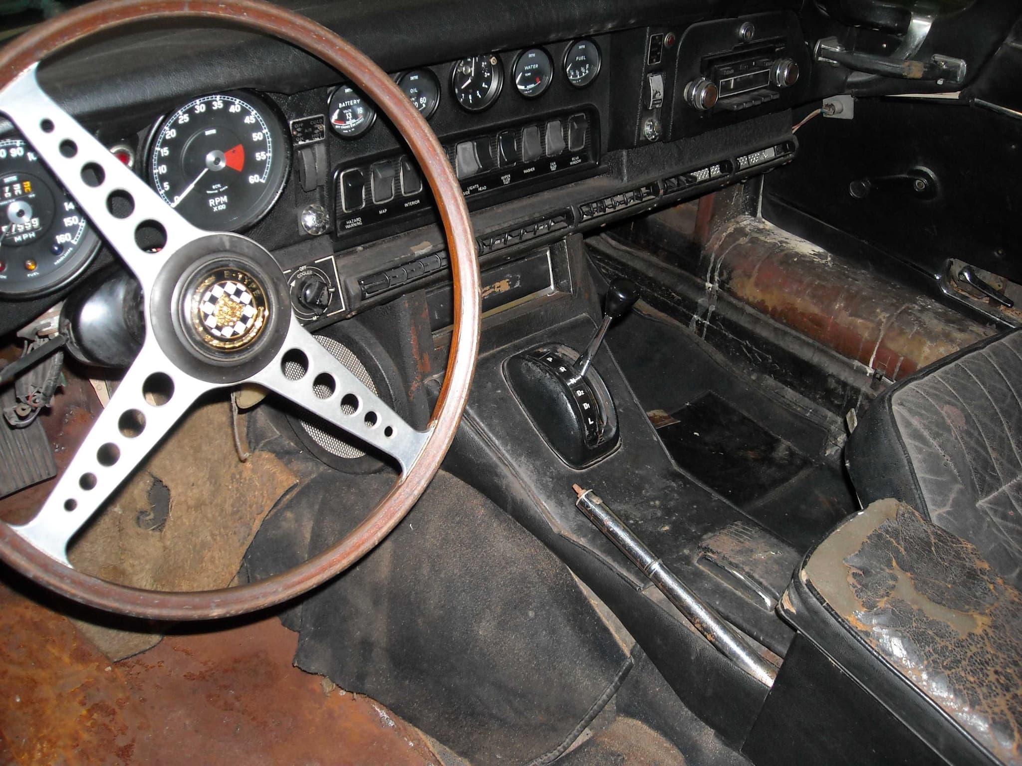 A neglected E-Type cockpit with rusty floor, rotten carpet and cracked leather around an automatic selector.