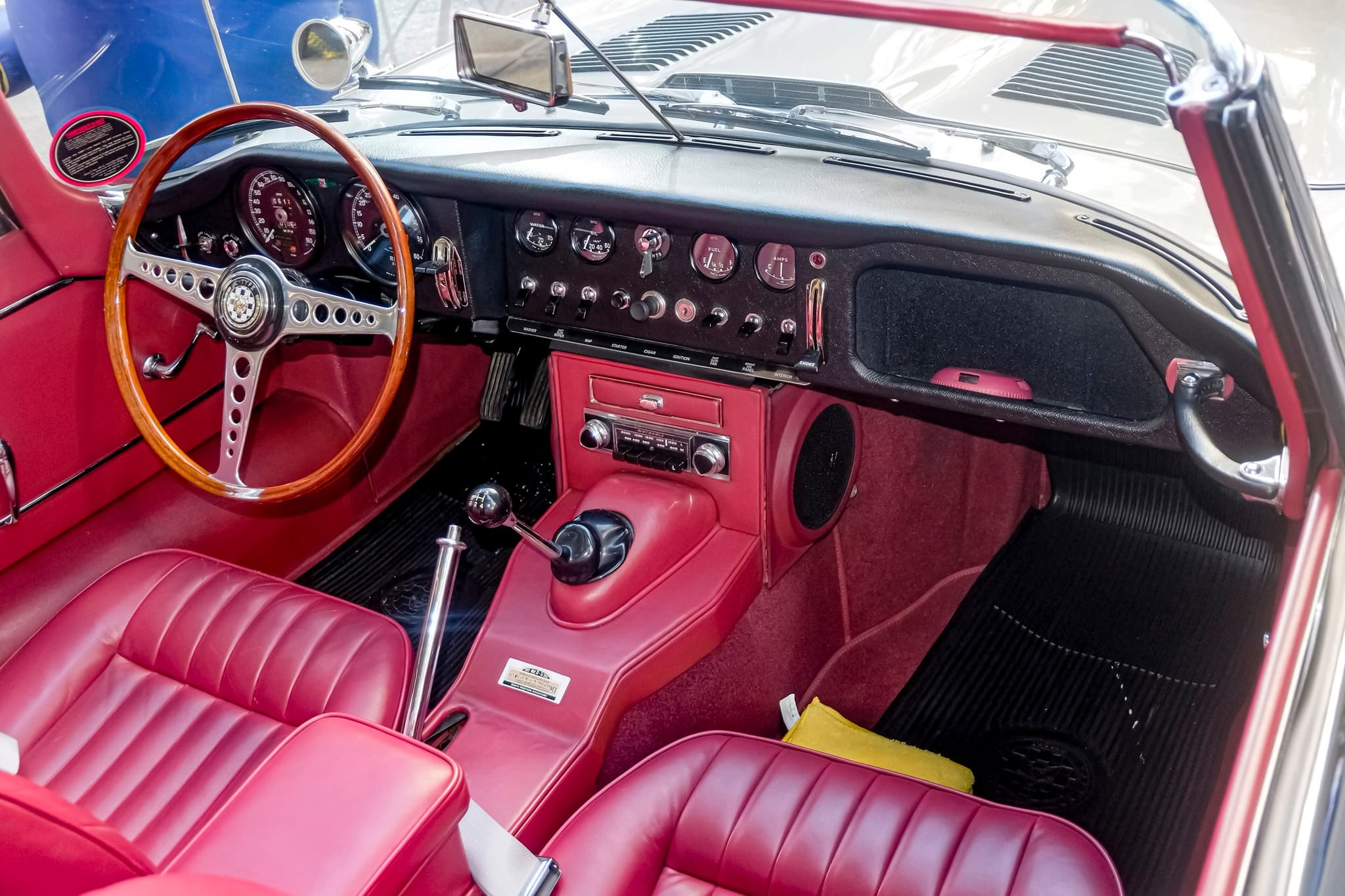 An E-Type roadster's cockpit with red leather seats, a black dash panel with toggle switches and a wood-rim wheel on the left.