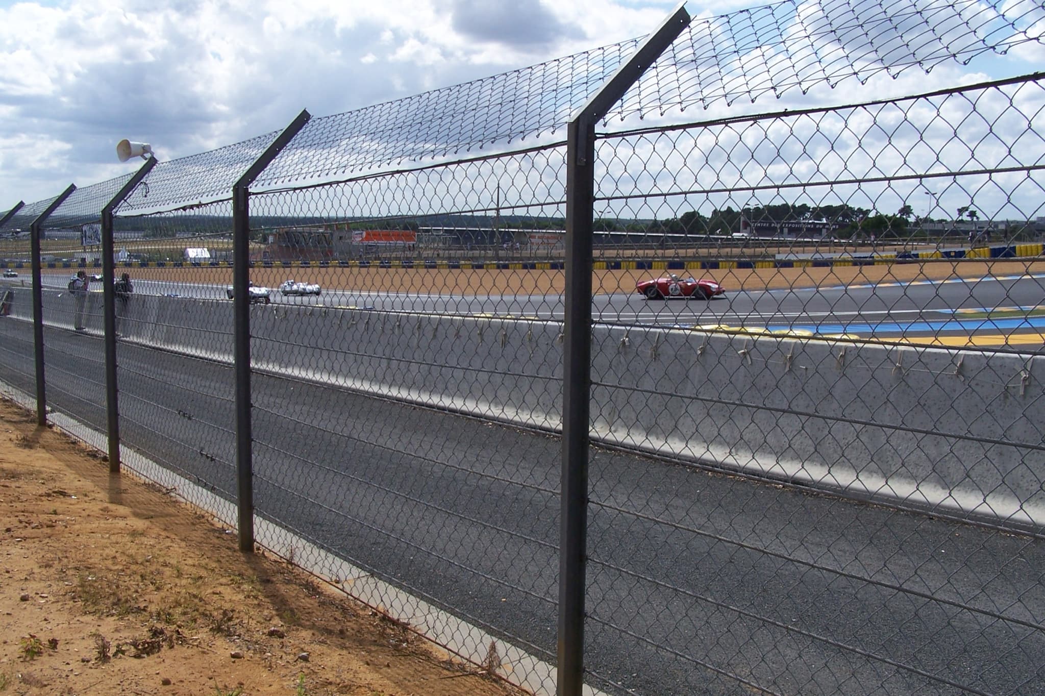 Racing cars far down a straight seen through a tall chain-link catch fence, with pits and grandstands beyond.