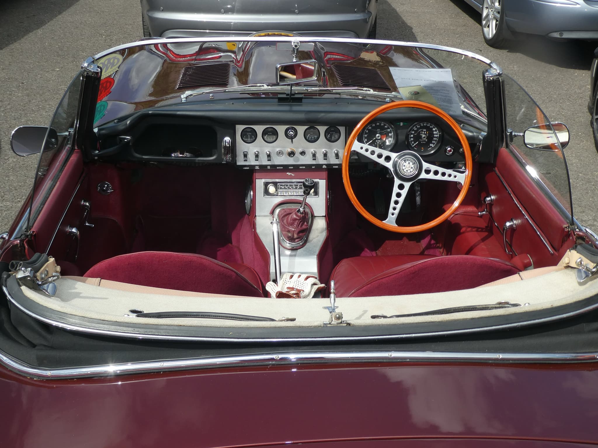 An early E-Type roadster's cockpit from behind, with red bucket seats, an aluminium console and a wood-rim wheel on the right.